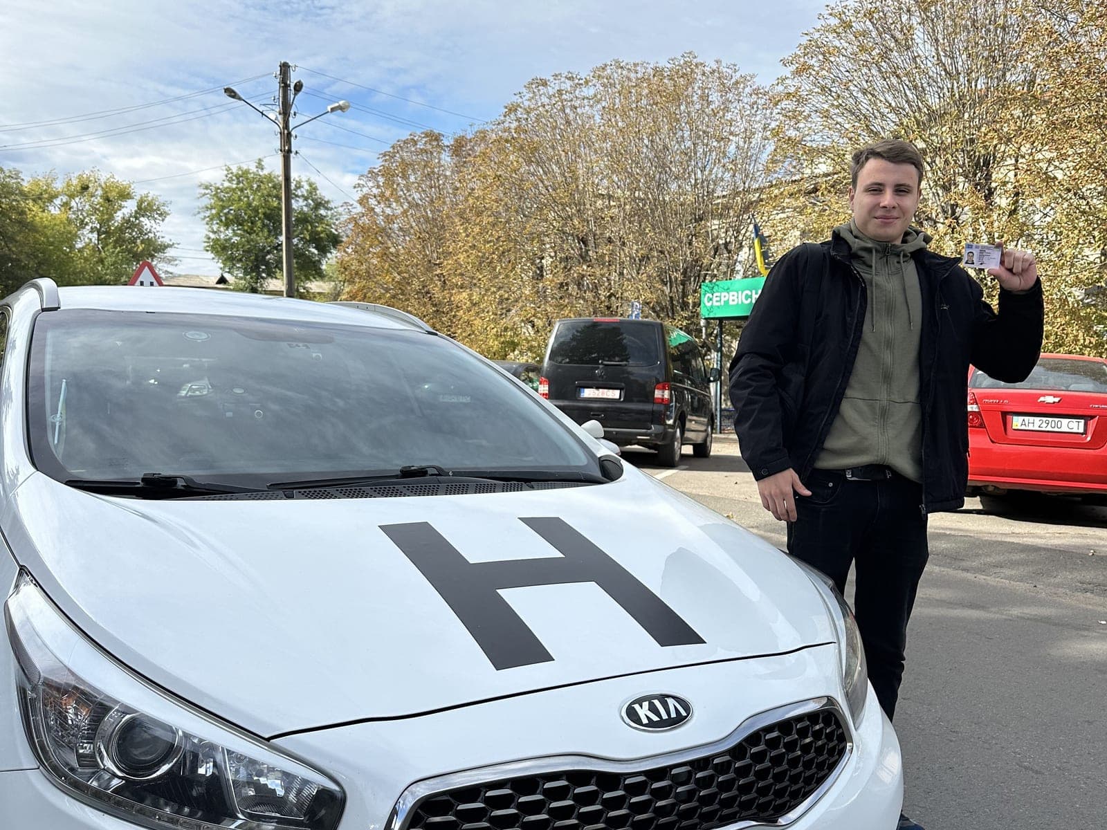 Випускник автошколи Лідер з посвідченням водія біля навчальної Kia зі знаком «Н»